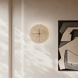 Eine elegante Wandlampe und Uhr Brecht, die minimalistisch in einem modernen Raum präsentiert wird. Die runde Uhr besteht aus einem natürlichen, steinähnlichen Material und verfügt über schlichte Zeiger in Gold. Sie strahlt ein warmes, indirektes Licht aus, das die Wand stilvoll beleuchtet. Ein zeitloses Wohnaccessoire für moderne Innenräume.
