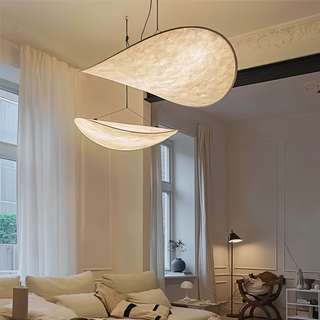 Zwei elegante, energieeffiziente LED-Hängelampen in Form von stilisierten Tropfen hängen an der Decke eines modernen Wohnzimmers. Die Lampen haben eine weiche, warme Ausstrahlung und ergänzen die minimalistische Einrichtung des Raums. Der Raum ist hell erleuchtet, mit weißen Vorhängen und einem gemütlichen Sofa im Vordergrund, was die beruhigende Atmosphäre verstärkt. Die Lampen bieten sowohl funktionale Beleuchtung als auch ästhetischen Charme.